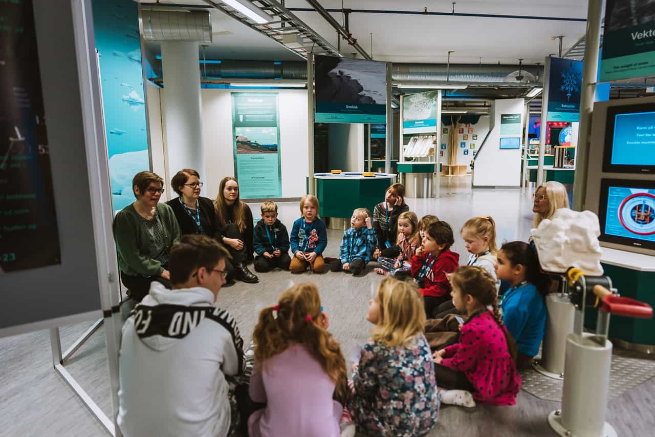 Arctic Frontiers - Children sit in a circle on the floor with adults in a science museum or educational centre, exploring Arctic Frontiers - Young activities and engaging in fun Science for Kids sessions.