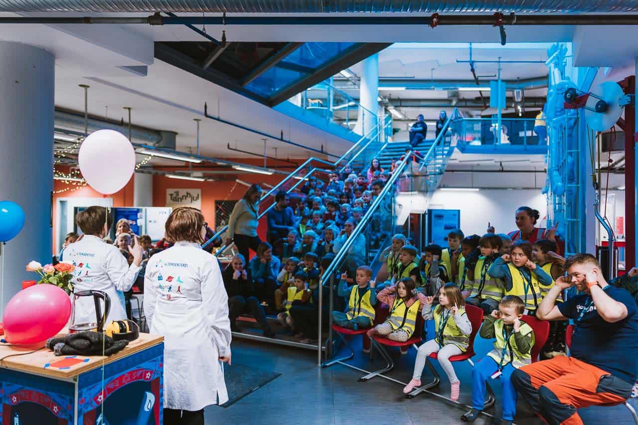 Arctic Frontiers - Children in yellow vests watch an Arctic Frontiers – Young science for kids demonstration indoors, with presenters in lab coats at the front.