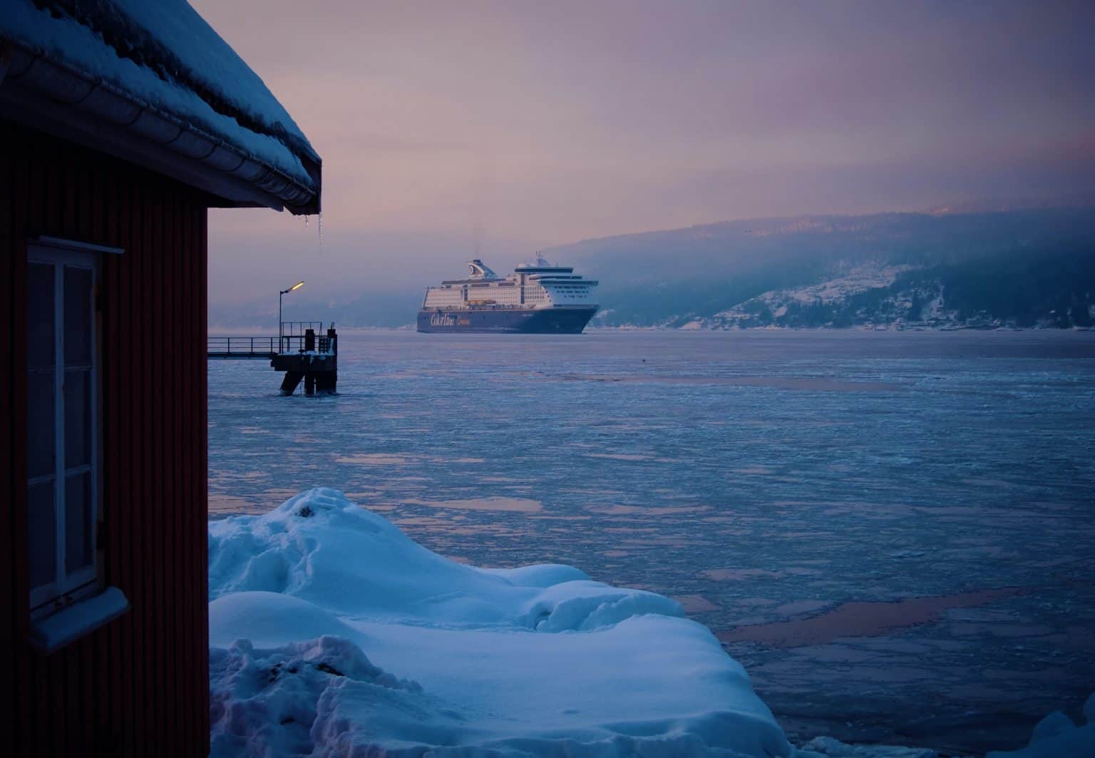Arctic Frontiers - A large cruise ship sails on an icy fjord at dusk, with snow-covered shore and a wooden cabin in the foreground, capturing the serene beauty of Arctic Frontiers.