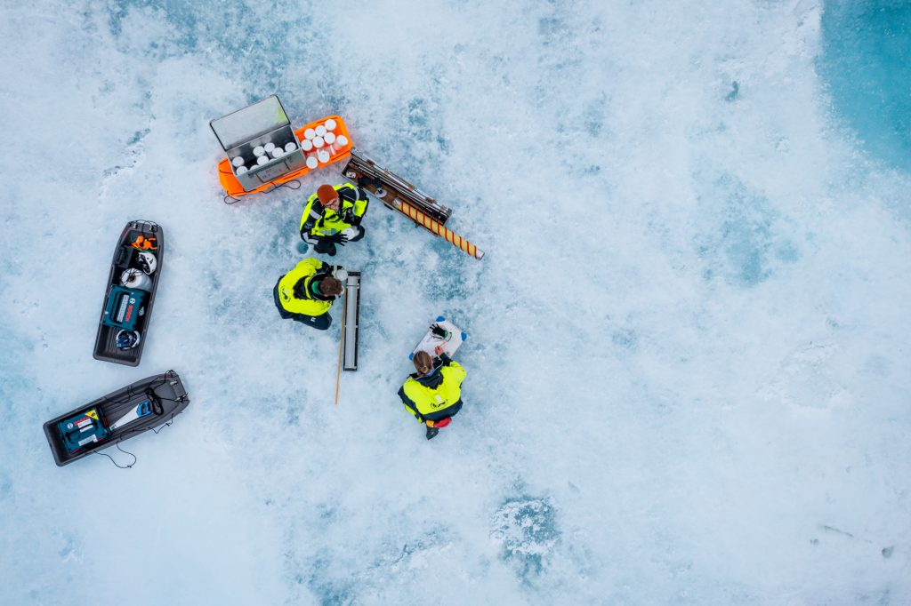 Arctic Frontiers - Aerial view of scientists in yellow jackets working with equipment on a snowy, icy surface, captured as part of the Arctic Frontier Partner pictures.
