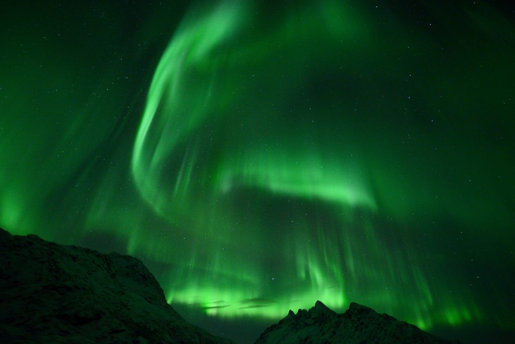 Arctic Frontiers - Green northern lights swirl above dark, snowy mountains under a starry night sky, capturing the inspiring spirit of Arctic Frontiers - Emerging Leaders - 2022 and older.