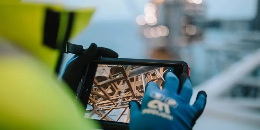 Arctic Frontiers - An Arctic Frontier Partner wearing blue gloves and a yellow jacket uses a tablet to take photographs of industrial metal beams.