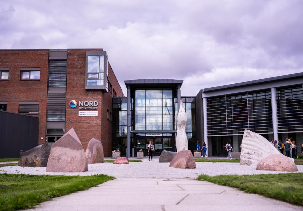 Arctic Frontiers - People walk near the modern brick and glass building of Nord University, an Arctic Frontier Partner, with stone sculptures in front.