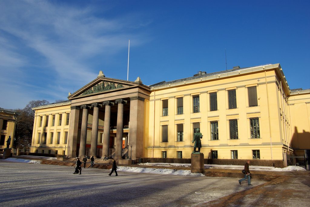 Arctic Frontiers - Yellow neoclassical building with columns, statue in front, people walking, and snow on the ground under blue sky—an iconic scene captured in Arctic Frontier Partner pictures.