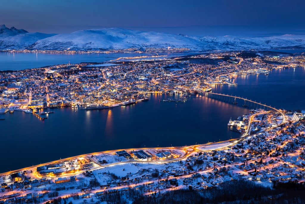 Arctic Frontiers - Aerial view of a snowy coastal city at dusk, illuminated by streetlights and surrounded by mountains—an inspiring scene perfect for Arctic Frontier Partner pictures.