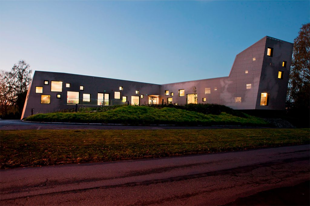 Arctic Frontiers - Modern building with uneven geometry and lit square windows, viewed at dusk with a grassy area in front—an architectural vision reminiscent of Arctic Frontier Partner pictures.