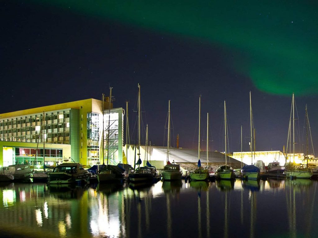 Arctic Frontiers - Boats moored at a marina at night with green northern lights in the sky and a modern building nearby, captured in stunning Arctic Frontier Partner pictures.