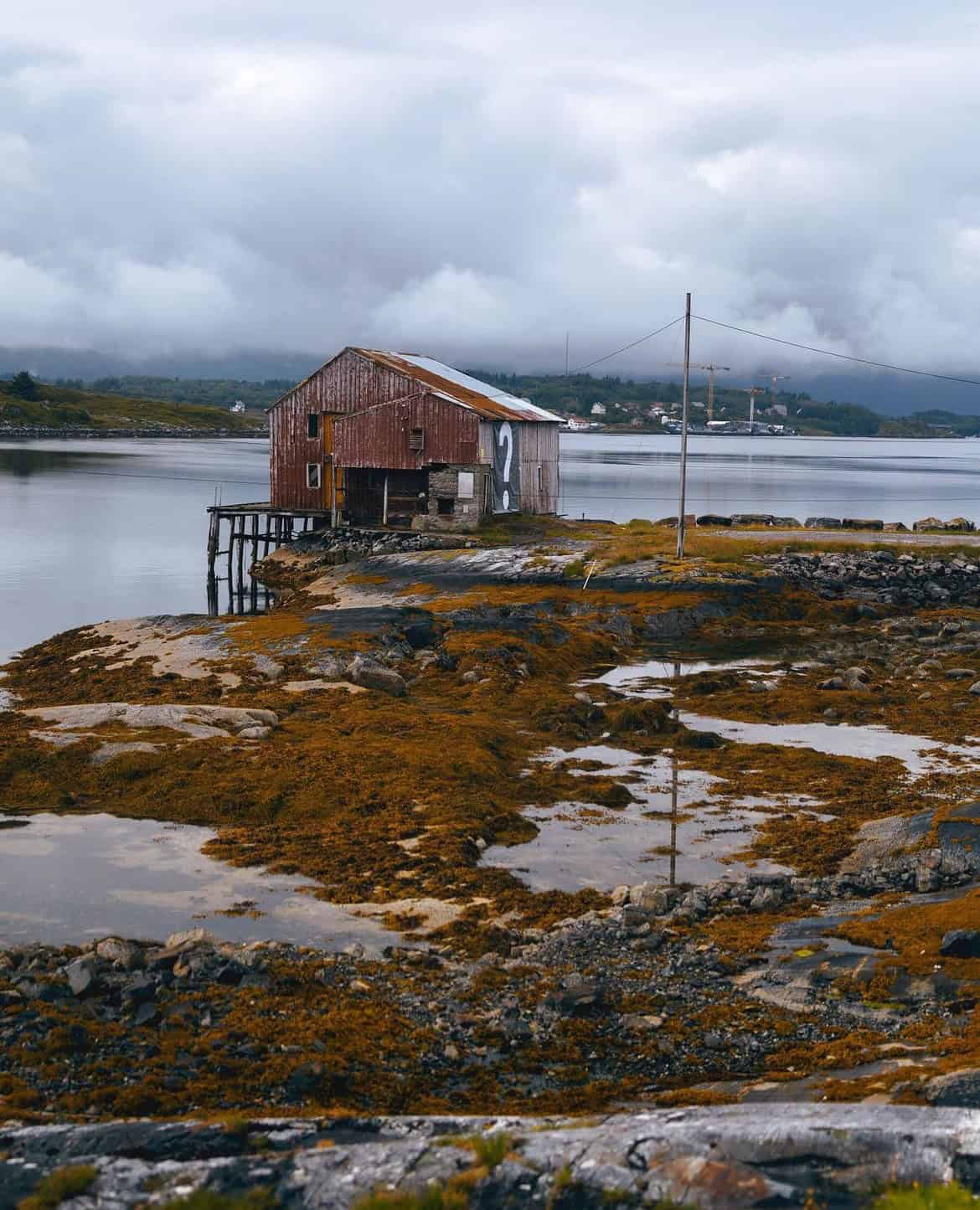 Arctic Frontiers - Old red wooden shed on stilts by a calm lake, surrounded by rocks and moss under a cloudy sky—evoking the quiet isolation of Arctic Frontiers.