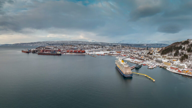 Arctic Frontiers - Aerial view of a snowy port city during Arctic Frontiers Open – Narvik, with ships berthed and mountains in the background under a cloudy sky.