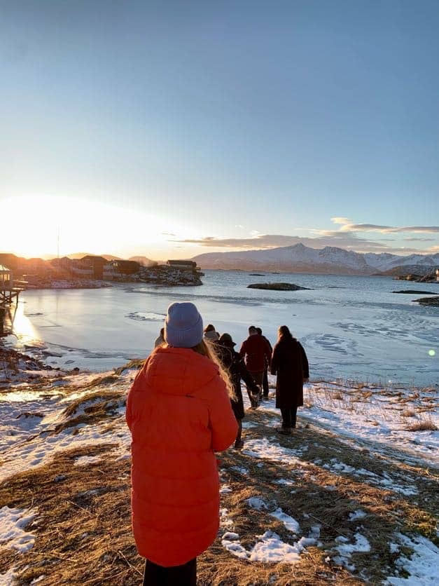 Arctic Frontiers - People in winter coats walk towards a frozen lake at sunset with snowy mountains in the distance, capturing the spirit of Arctic Frontiers - Kirkeneskonferansen.