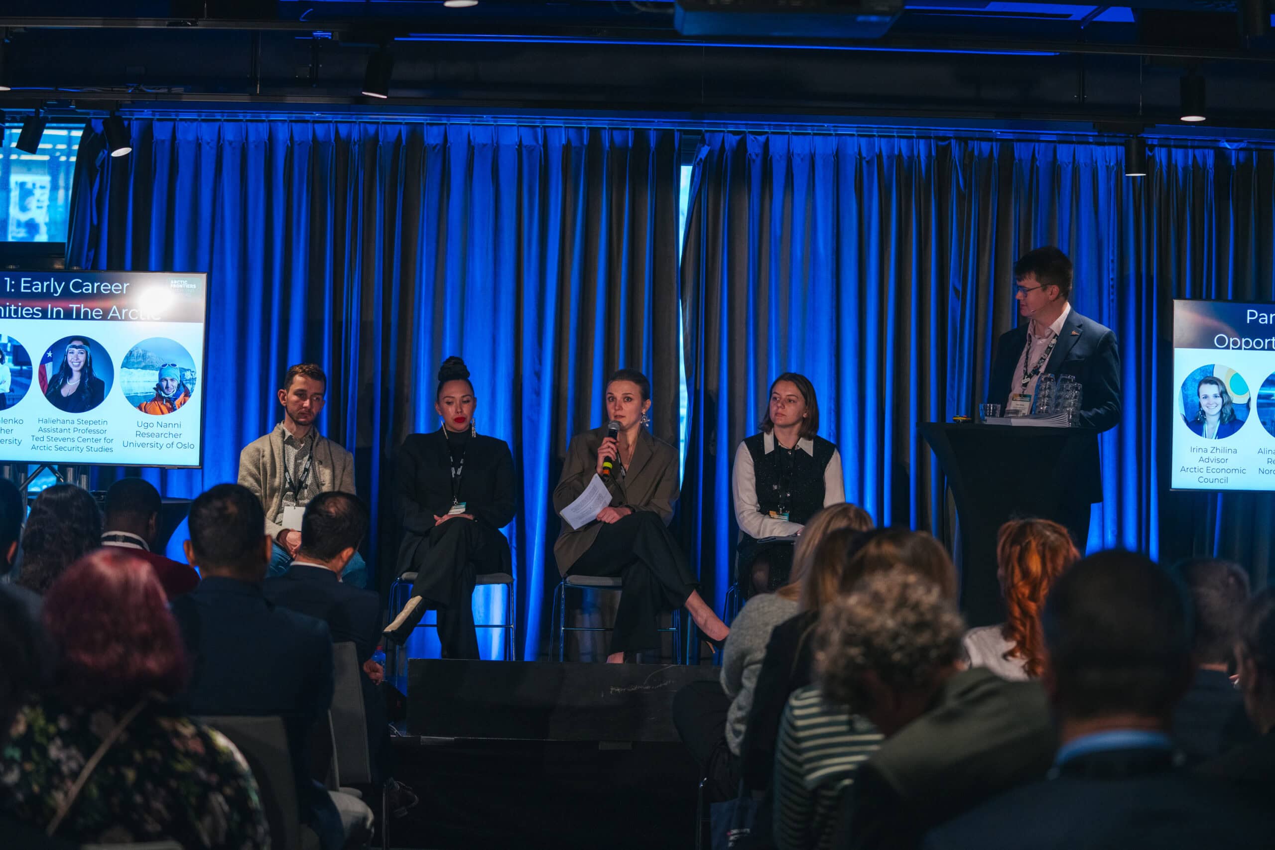 Arctic Frontiers - Five people on stage at an Arctic Frontiers – Emerging Leaders Alumni Network panel discussion, engaging with an attentive audience in a conference room bathed in blue lighting.