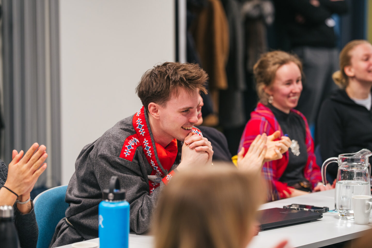 Arctic Frontiers - Smiling young people sit at a table, applauding and chatting during the Arctic Frontiers Student Forum group meeting or event.