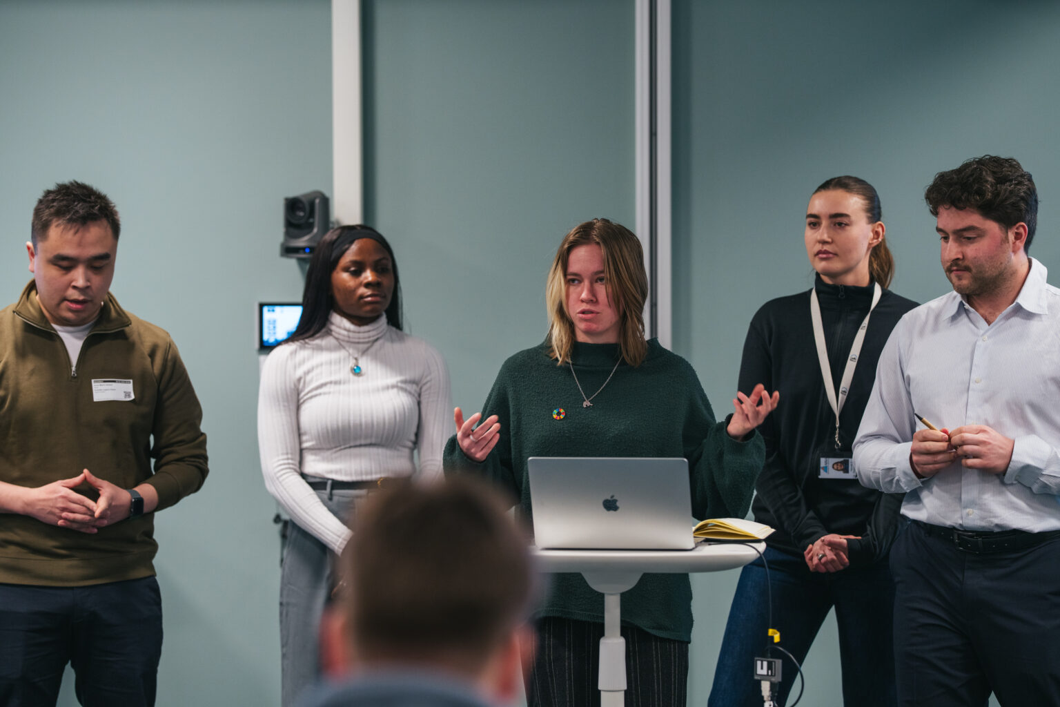 Arctic Frontiers - Five people stand at the front of a room; one woman speaks while others listen near a laptop on a lectern during the Arctic Frontiers Young Student Forum.