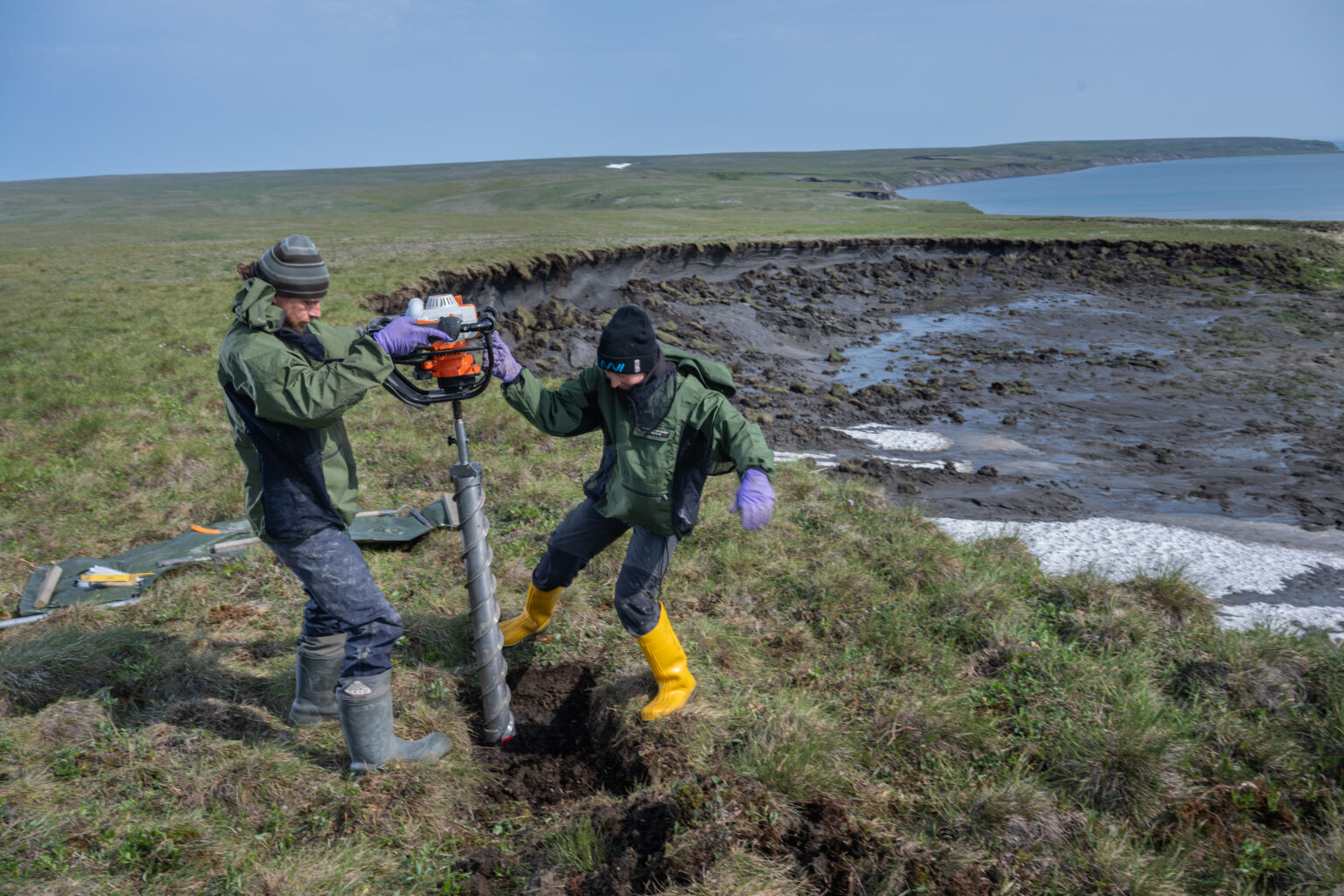 Arctic Frontiers - Two people in outdoor gear drill into grassy ground near a muddy, eroded landscape with water in the background, gathering data for the Arctic Frontiers - Science 2026 conference.