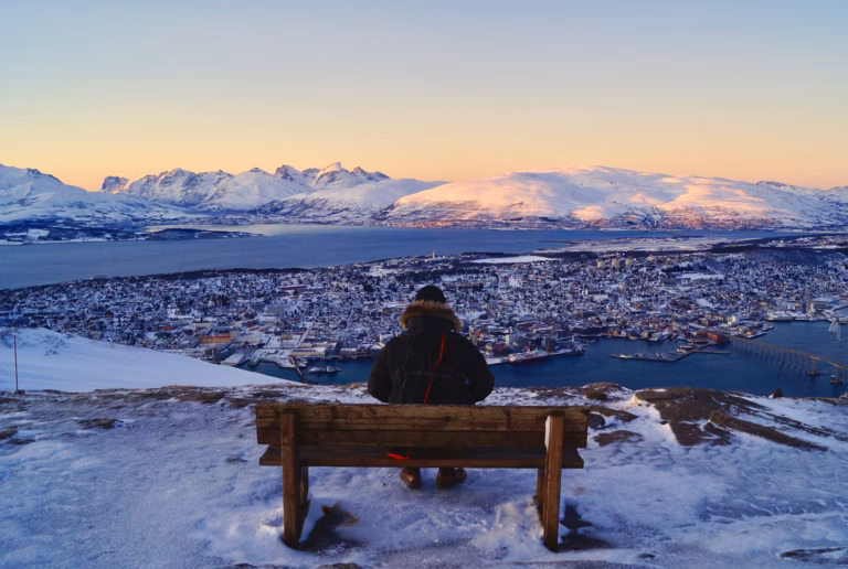 Arctic Frontiers - A person sits on a bench overlooking a snowy EU cityscape and distant mountains at sunset.