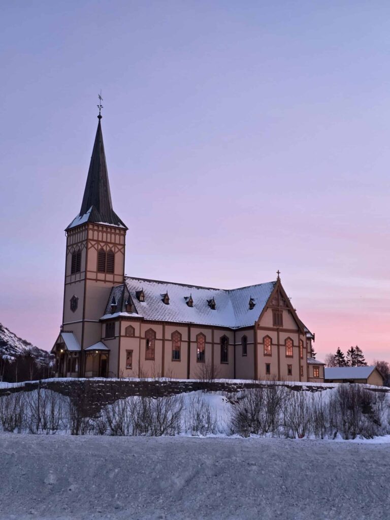 Arctic Frontiers - A snow-covered church with lit windows at sunset, under a purple and pink sky.