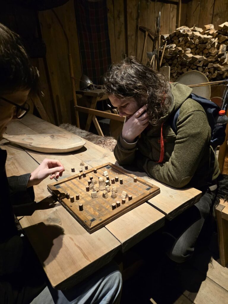 Arctic Frontiers - Two people sit at a wooden table, focused on playing a board game with wooden pieces in a rustic room.
