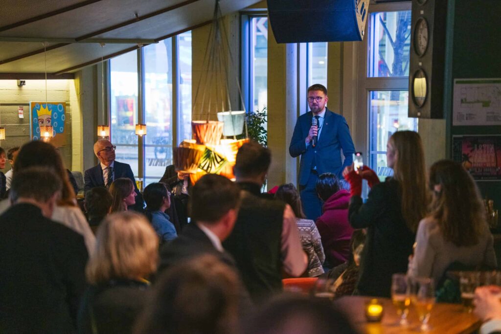 Arctic Frontiers - A man in a suit speaks to a seated crowd in a warmly lit indoor event space.