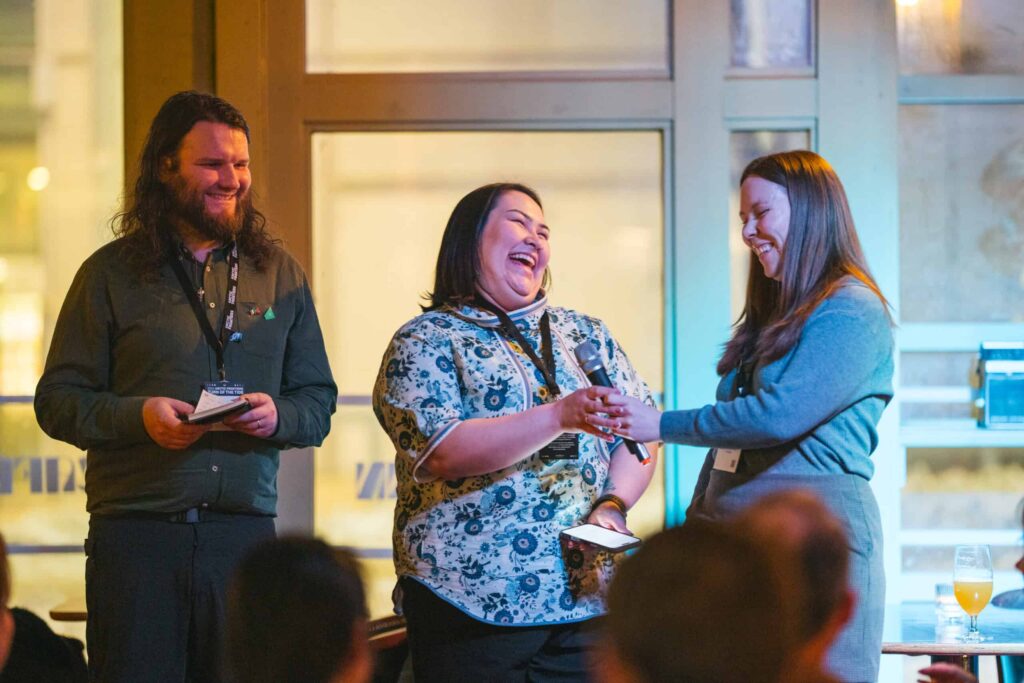 Arctic Frontiers - Three people on stage laugh and smile while one hands a microphone to another at an indoor event.