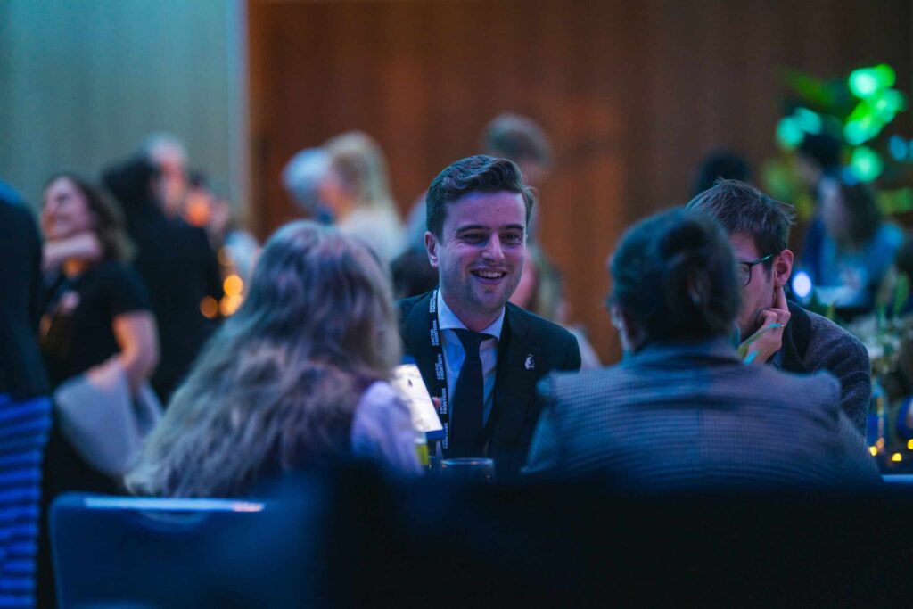 Arctic Frontiers - A man in a suit smiles whilst talking to others at a lively indoor event with dim blue lighting.