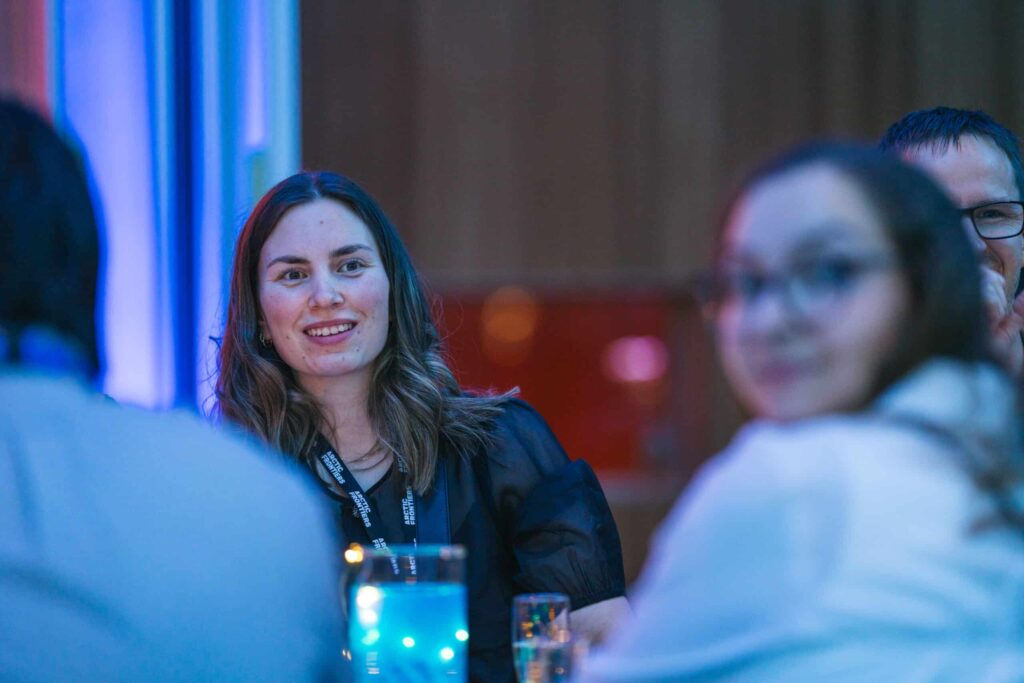 Arctic Frontiers - A woman with brown hair smiles at a table during an indoor event with blue lighting.