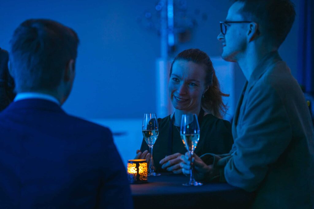 Arctic Frontiers - Three people in semi-formal attire chat and enjoy drinks by candlelight at a blue-lit event.