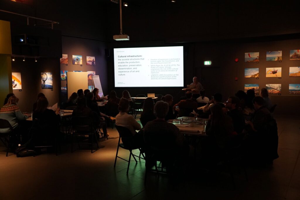 Arctic Frontiers - A group of people sit in a dim room, watching a bright presentation projected onto a screen.