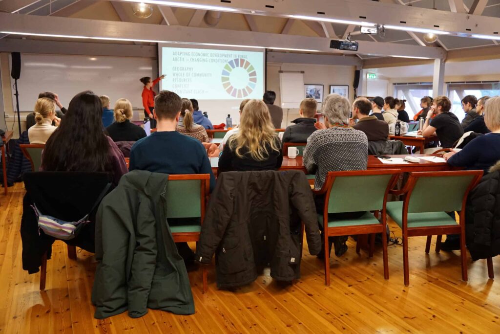 Arctic Frontiers - People sit at tables facing a presentation screen in a well-lit room with wooden floors and exposed beams.
