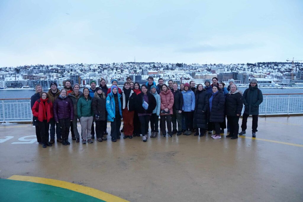 Arctic Frontiers - A group of people poses outdoors on a decking with a snowy town and water visible in the background.