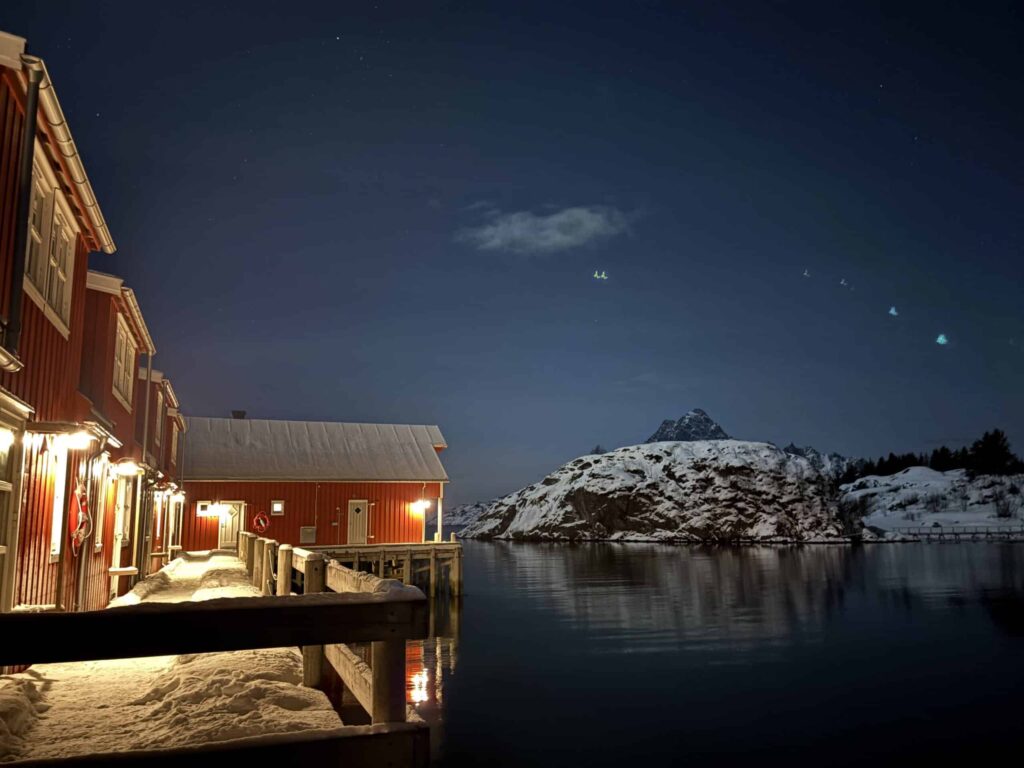 Arctic Frontiers - Red cabins with warm lights reflect on calm water, snowy mountain in the background under a night sky.